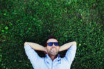 Photo of a man lying in the grass with sunglasses on and his arms behind his head