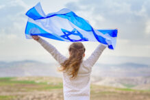 Stock image of a woman facing away from the camera holding up an Israeli flag - desert scenery in background.
