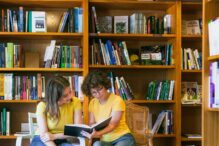 photo of two teens wearing yellow shirts and jeans sitting in front of a bookshelf stacked with books holding a book and reading together