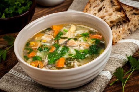 photo of a bowl of chicken soup filled with carrots, chicken, celery, herbs and broth on top of a napkin with two pieces of hearty bread