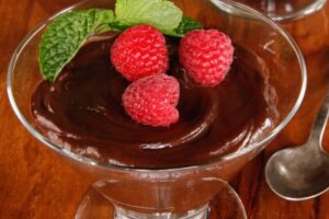 photo of vegan dark chocolate mousse in a glass dish topped with raspberries and mint sitting on wood table with a spoon