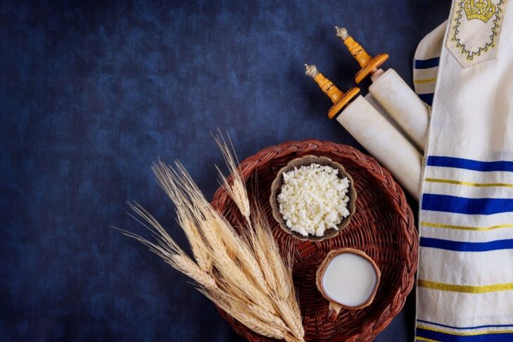 photo of a small Torah covered by a tallit next to a woven basket with wheat stalks, cheese and milk