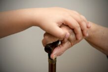 photo of a child's hand on top of an elderly person's hand that is holding a walking cane