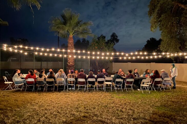 Image of a group of people sitting at a long communal table outside.