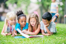 photo of four kids reading a book together on the grass