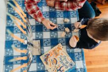 photo of a parent helping a kid design a wooden dreidel with extra dreidels laying on the table next to a Hanukkah book and a menorah filled with unlit candles