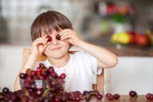 photo of a young child holding cherries over their eyes while sitting on a chair with a large bowl of cherries overflowing on the table in front of them