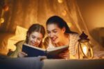 photo of a mother and daughter reading a book together at night by a lamp
