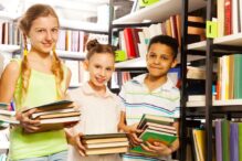 photo of three kids holding stacks of books standing next to library shelves