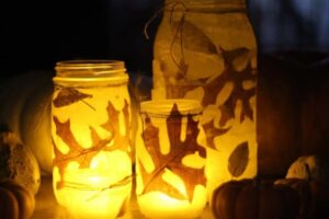 photo of three handmade votives decorated with leaves and fall colors and lit up by candles next to some gourds