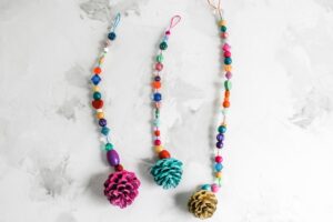 photo of three hanging decorations including a variety of beads and a painted, glitter pinecone laying on top of a marbled surface