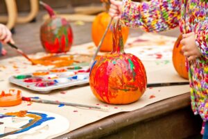 photo of kids painting pumpkins on a table lined with paper and filled with paint and materials