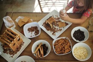 photo of a table filled with ingredients to make an edible sukkah including pretzels, nuts, chocolate, cinnamon sticks and graham crackers while a little girl is squeezing frosting onto a graham cracker