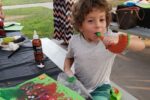 photo of a boy blowing a toy shofar at a park sitting in front of an art project