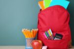 Stock image of a red backpack filled with colorful notebooks, pens and a calculator sitting next to a pencil holder with sharpened pencils and an apple
