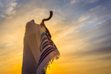 Image of someone blowing a shofar at dusk while wearing a tallit.