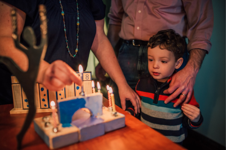 Image of a small child standing near a menorah as his parents light it