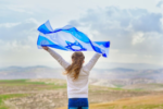 Girl holding Israeli flag blowing in the wind facing away from the camera toward the land of Israel